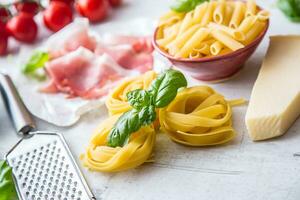 Italian or mediterranean food cuisine and ingredients on white concrete table. Tagliatelle pene pasta  olives olive oil tomatoes parmesan cheese prosciutto and basil leaves on white concrete board photo