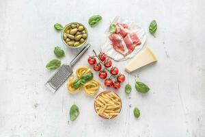 Italian or mediterranean food cuisine and ingredients on white concrete table. Tagliatelle pene pasta  olives olive oil tomatoes parmesan cheese prosciutto and basil leaves on white concrete board photo