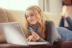 hermosa joven niña participación crédito tarjeta comprando en línea a sofá en vivo habitación foto