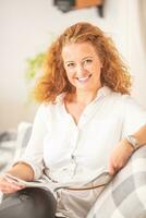 Woman with curly hair reads a magazine, sitting on a sofa in a living room, smiling at the camera photo