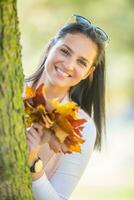 Happiness woman sitting in autumn park holding bouquet with fall leaves cute look from behind a tree photo