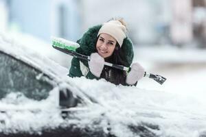 Smiling well clothed woman holds ice scraper and snow broom while leaning against the car covered in snow photo