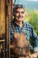 Older and experienced craftsman stands smiling in front of the workshop in checkered shirt and leather apron photo