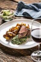 Red wine as a typical baverage to a red meat steak with roasted potatoes, spinach dip and fresh salad served on a white porcelain plate on the wooden desk. photo