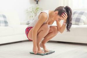 Young woman squats on a scale holding her head frsutrated over gaining weight photo