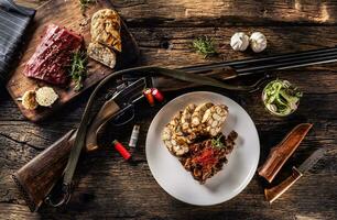 Rustic wooden table with a raw deer venison, delicious homemade dumpling and rosemary. Between the plate full of venison goulash and dumpling lies a hunting gun with bullets and a knife photo