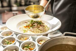 Chef in restaurant kitchen pours broth into a chicken soup with noodles, meat and vegetable photo
