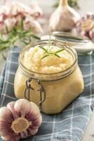 Aromatic garlic paste in a glass jar laid on rustic kitchen cloth with bulbs and peeled cloves and rosemary photo