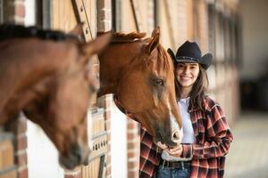 Good-looking girl in cowgirl hat stands next to horses inside the stable, smiling photo
