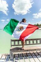 Woman holds Mexican flag behind her back as she stands on a bench on a pier next to the Carribean Sea photo