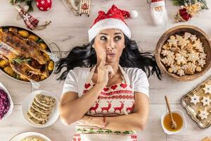 Pensive female cook in a Christmas apron and Santa hat,l ying on the floor, surrounded by gingerbread, Linz cakes, on the other side, roast goose or turkey with side dishes looks to the side photo