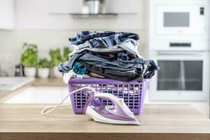 Big pile of wrinkled clothes pin a purple basket on a kitchen desk with an iron in front of it photo