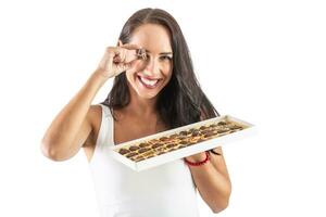 Happy brunette is smiling widely with candy box in her hand and a little chocolate in fron of her eye. Isolated background photo