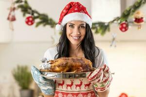 Satisfied young woman in a Christmas apron and Santa hat holding a roasting pan with a roast goose or turkey photo