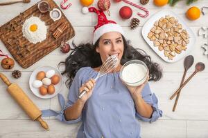 Good-looking dark haired girl tasting creme on stirrer and laying on the ground and being surrounded by gingerbreads, eggs, flour on a wooden desk, christmas hat, dried oranges and baking forms photo