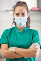 Young good-looking female healthcare worker in green wears face mask and protective gloves. standing with arms crossed photo