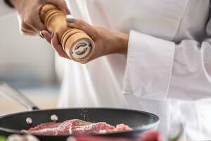 Detail of a wooden pepper grinder putting spice on cut of pork in a pan photo