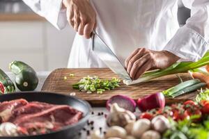 Detail of a person cutting spring onion on a copping board with raw meat and vegetables ingredients around photo