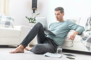 Male freelancer sits barefoot on the living room floor working on a laptop photo