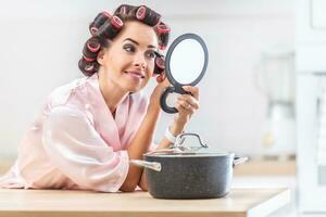 Housewife doing her makeup looking into the mirror leaning against the kitchen desk with a pot next to her photo