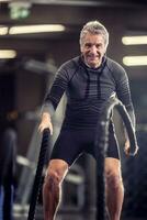 Older man with headphones exercises with ropes in a fitness training center photo