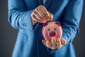 Saving money. Businesman holding pink piggy  and putting coin into piggy bank. photo