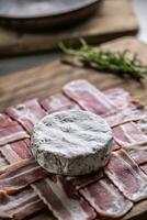 Brie cheese disk on a zigzag slices of bacon during food preparation on a wooden plate photo