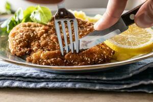 Fork and knife taking a first cut from a schnitzel in sesame seeds served with a slice of lemon photo