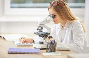 Beautiful young researcher looking into a microscope, writing down notes photo