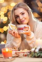 un joven niña en un lana suéter participación un pan de jengibre y un taza con té café o Navidad puñetazo foto