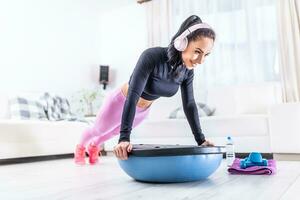 ajuste guapo hembra usa auriculares durante su hogar rutina de ejercicio participación tablón posición con manos en un equilibrar pelota foto