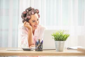 Female in hair rollers and nighgown laughs at notebook scrren while sittinf at the table at home photo