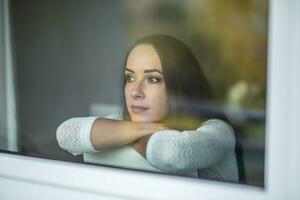 Sad good-looking female sits on a chair behind the window looking outside photo