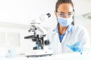 Female lab technician in protective glasses, gloves and face mask sits next to a microscope and conical flask, looking aside on the desk photo