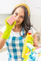 Sexy young woman smiling into the camera, holding a dish washing sponge in her rubber gloves, partially covering her face. Holding detergent in the other hand with more cleaning stuff in front of her photo