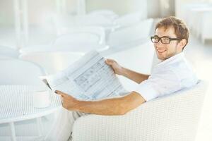 un hombre en lentes leyendo un periódico mientras sentado a un mesa foto