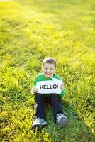 a young boy sitting in the grass holding a hello sign photo