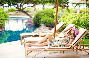 a woman in a floral dress and hat is relaxing on a lounge chair near a pool photo