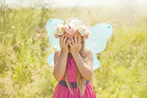un pequeño niña en un rosado vestir con mariposa alas cubierta su ojos foto