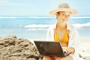 a woman in a hat and bikini sitting on the beach with a laptop photo