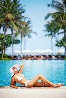un mujer en un blanco sombrero y bikini tendido en el borde de un piscina foto