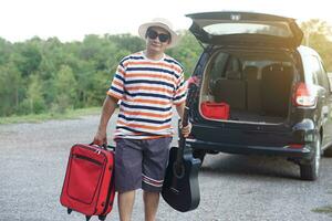 asiático hombre viajero va de viaje por auto, sostiene rojo maleta y el guitarra. concepto, relajarse tiempo. vacaciones, fiesta o fin de semana viaje por privado vehículo. transporte. la carretera viaje.regalar hora para tú mismo foto