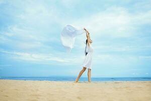 un mujer en un blanco vestir en pie en el playa con un blanco paño foto