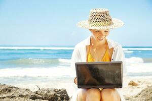 a woman in a hat and bikini sitting on the beach with a laptop photo