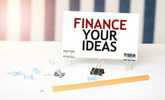 FINANCE YOUR IDEAS sign on paper on white desk with office tools. Blue and white background photo