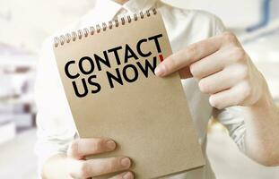 Text CONTACT US NOW on brown paper notepad in businessman hands in office. Business concept photo