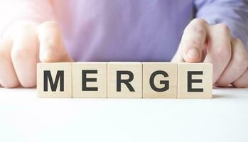 Businessman hand holding wooden cube block with MERGE business word on table background. photo