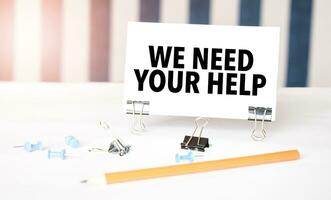 we need your help sign on paper on white desk with office tools. Blue and white background photo