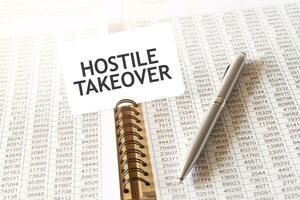 Text HOSTILE TAKEOVER on paper card, pen, financial documentation on table photo