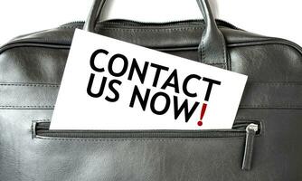 Text CONTACT US NOW writing on white paper sheet in the black business bag. Business concept photo